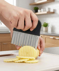Side view of a stainless steel crinkle cutter with a black handle slicing a potato on a cutting board.