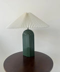 Green ceramic table lamp with pleated white fabric shade on wooden table.