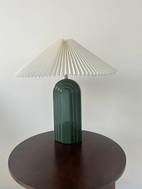 Green ceramic table lamp with pleated white fabric shade on wooden table.