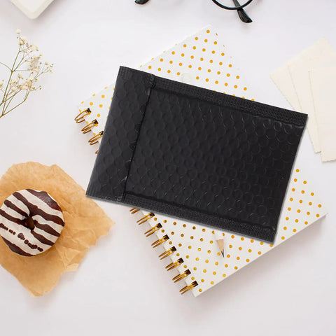 Black padded bubble mailer next to polka dot notebooks and a donut, part of 100 pack, 12x18 padded bubble mailers self seal envelopes.