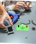 Hands assembling Mini Solar Powered DC Motor For Science Experiments And Hands On DIY Projects with tools and parts on table.