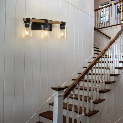 Farmhouse 3 Light Vanity Fixture With Rustic Wood And Clear Glass Shades for hallway, mounted on white paneled wall.