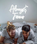 Couple in bed with "Always & Forever" white LED neon sign on wall, similar to Lets Celebrate Neon Sign White Letter LED Neon Light For Wall Decor USB Powered Word Light For Party Bar Club Bedroom Living Room Hotel Gift 16.9x10.2 Inches