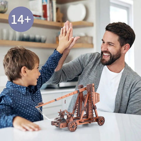 Father and son high-five next to DIY 3D Wooden Trebuchet Puzzle Kit for Adults Hands On Catapult Engineering Toy on table.