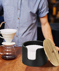 Coffee Filter Holder And Countertop Storage Jar With Lid For Easy Organization, wooden with filters next to pour-over coffee setup.