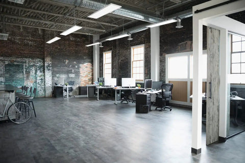 Modern office space with desks and computers under exposed ductwork ceiling with hanging lights.