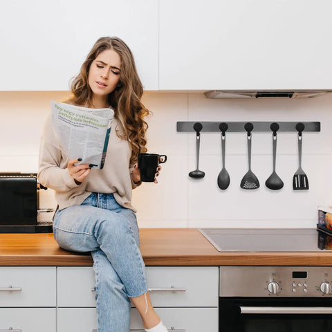 Woman sitting on kitchen counter reading newspaper with utensils hanging on wall rack in modern kitchen setting.