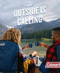 Two people in camping chairs by a lake with mugs, surrounded by mountains and forest. Coleman logo on chair. Text reads 'The Outside is Calling.'