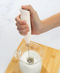 Powerful Handheld Milk Frother For Lattes And Cappuccinos Battery Powered Mini Drink Mixer Perfect For Home Barista Use Creamy Foam In Seconds Great For Matcha And More being used to froth milk in a glass jug on a wooden board.