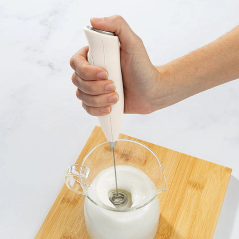 Powerful Handheld Milk Frother For Lattes And Cappuccinos Battery Powered Mini Drink Mixer Perfect For Home Barista Use Creamy Foam In Seconds Great For Matcha And More being used to froth milk in a glass jug on a wooden board.