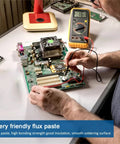 Person applying No Clean Rosin Solder Flux Paste For Electronics on circuit board, 100g pack of 2, with multimeter nearby.