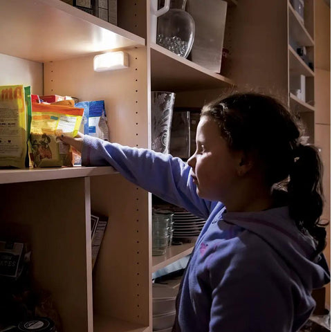 Girl reaching for snacks in a dimly lit pantry illuminated by a Wireless Motion Sensing LED Night Lights Six Pack Tiny Stick Anywhere White Battery Powered Auto On When Motion Is Detected In Darkness Great For Hallways Closets And Stairs.