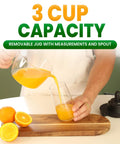 Primtronic Electric Citrus Juicer With Large 24 Oz Capacity And Seed Filter Auto Reverse Function Compact Design For Oranges Lemons Limes BPA Free Black Orange pouring fresh orange juice from a 3-cup capacity pitcher into a glass.