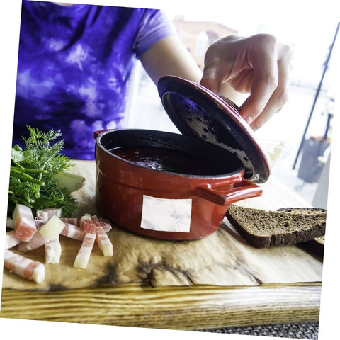 Person lifting lid of a red ceramic pot beside sliced bread and herbs on a wooden surface.