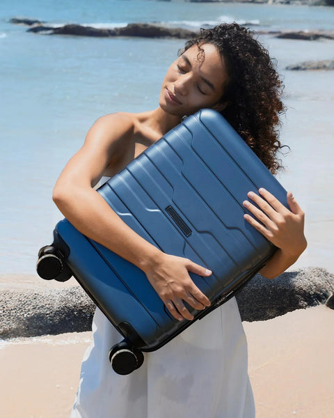 BagSmart blue hard-shell carry-on luggage with spinner wheels, held by a woman at the beach