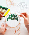 Person embroidering cactus design in hoop with thread; various colored threads and tools on table