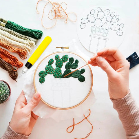 Person embroidering cactus design in hoop with thread; various colored threads and tools on table