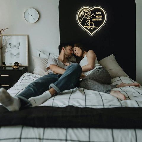 Couple in bed under a heart-shaped neon sign with wedding anniversary message, bedroom decor.