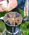 Bold Black Nine Cup Stovetop Espresso Maker Moka Pot brewing coffee outdoors in green setting, with aluminum body.