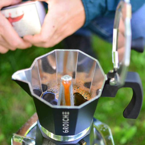 Bold Black Nine Cup Stovetop Espresso Maker Moka Pot brewing coffee outdoors in green setting, with aluminum body.
