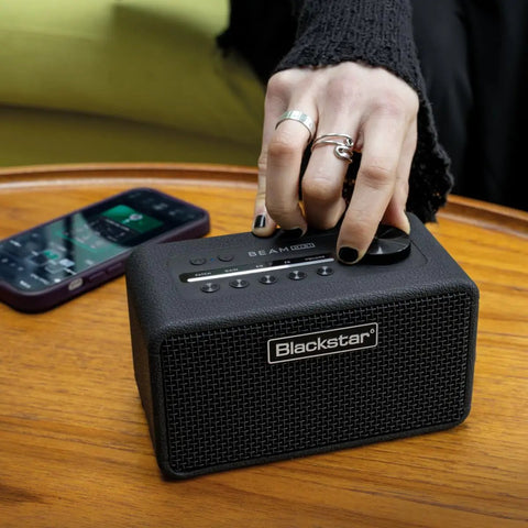 Person adjusting volume on Beam Mini Portable Guitar Amp With NAM Compatibility, 17 Models, 35 Plus Effects, USB C And Bluetooth, Dual 60 mm Full Range Drivers For Room Filling Stereo on wooden table.