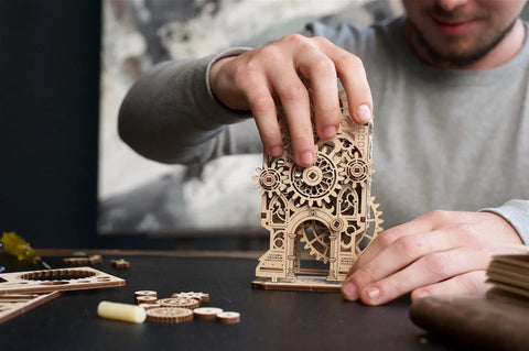Old Clock Tower 3D Wooden Puzzle Kit For Adults, DIY Mechanical Model Building Set, Wooden Clock Tower Project For Grownups, Brain Teaser Hobby, Relaxing Home Decor And Hands On Fun. Man assembles wooden gear clock tower model on table.