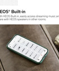 Smartphone displaying HEOS app interface on a brown couch, emphasizing HEOS built-in streaming feature for music control.