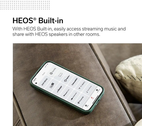 Smartphone displaying HEOS app interface on a brown couch, emphasizing HEOS built-in streaming feature for music control.