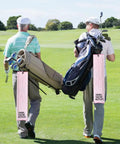 Two golfers walking with bags adorned with Swing Swear Repeat Embroidered Pink Golf Towel With Clip For Golf Bags Funny Ladies Golf Towel Perfect Gift For Moms Grandma Sister Aunt Golfer Retirement Birthday Mothers Day Sport Lover on a sunny course.