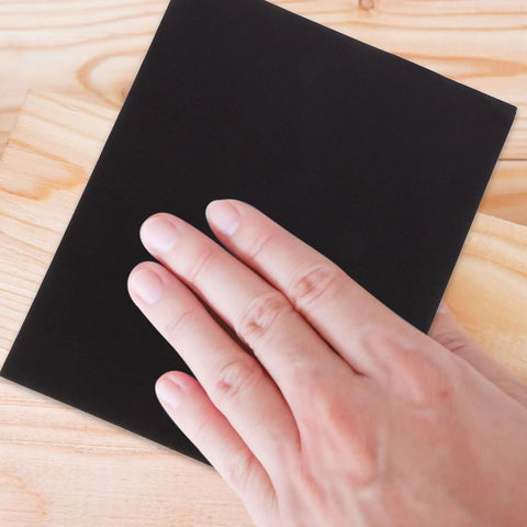 Person's hand touching black sanding sheet on wood surface from Top Tier 9 Sheets Wet Dry Sanding Paper Assortment featuring 9 Grits That Work Dope On Cars And Wood For Smoother Finishing And Polishing Like A Pro Without The Hassle Of Finding The Right Grit Each Time.