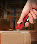 Auto Retracting Utility Knife With Dual Safety System For Fast Box Cutting Blade Auto Retracts When Off The Cutting Surface No Brainer Safety Push Button Release Easy Grip Handle Ideal For DIY Packing And Heavy Duty Work. Red metal utility knife cutting a sealed cardboard box in a warehouse