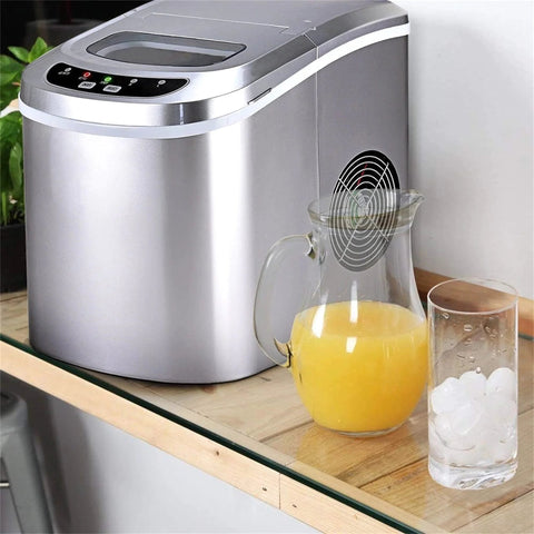 Front view of a stainless steel countertop ice maker with a digital control panel; juice pitcher and glass of ice nearby.