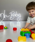 Child playing with toys in front of lit neon sign "Oh Baby" for wall decor, 31.5 by 15.7 inches, dimmable, cool white, for events.
