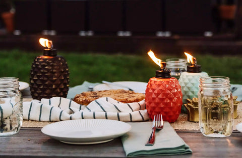 Colorful glass pineapple-shaped torches on outdoor dining table, demonstrating the 65 Inch Glass Pineapple Outdoor Torch With Copper Finish, Mosquito Repellent Torch For Yard And Garden, Integrated Snuffer, Burns Up To 15 Hours Per Fill, Use With Bite Fighter Torch Fuel, Patio Table Lighting That Stays Lit.