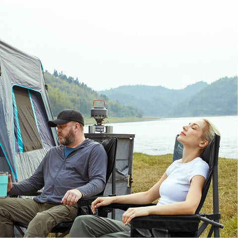 Two people relaxing in Oversized Folding Camping Chair With High Back Support 500 Lbs Capacity by a tent and lake.