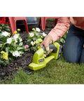 Person using ONE Plus 18 Volt Cordless Grass Shear And Shrubber for easy yard trimming near flowers, lightweight with overmold grip