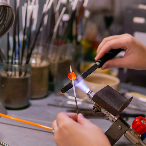 Craftsman using torch to shape glass bead in a jewelry workshop, with flame and tools visible on workbench.