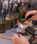 Person using a torch to melt a small orange glass piece for crafting in a workshop setting, surrounded by tools and materials.