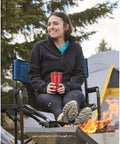 Person sitting on oversized tall director chair for camping and tailgating, heavy duty folding camp chair with 500 lb capacity, near campfire.