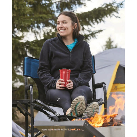 Person sitting on oversized tall director chair for camping and tailgating, heavy duty folding camp chair with 500 lb capacity, near campfire.