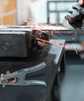 Heavy Duty Welding Ground Clamp For Mig Tig And Mma Welders with copper jaw grip on metal workbench amidst sparks.