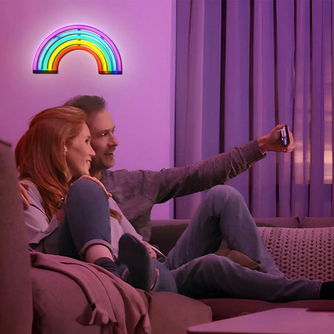 Couple on sofa using phone with Rainbow Neon Light Sign for Bedroom LED Colorful Rainbow Wall Decor on wall in background.