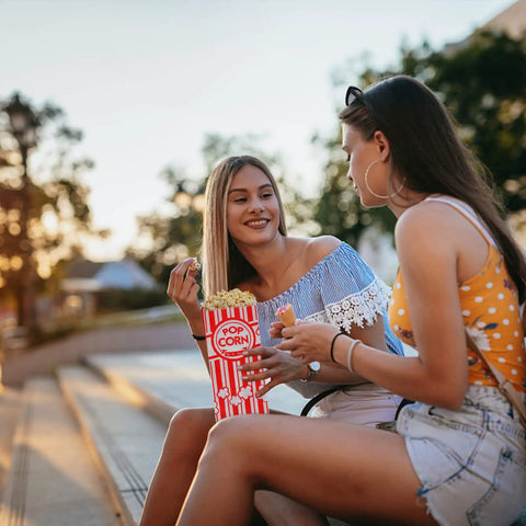 Two women sitting outdoors sharing popcorn from Heavy Duty Paper Popcorn Bags 1 Oz Individual Servings 500 Pack For Popcorn Machines