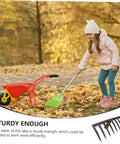 Child playing with a toy rake and wheelbarrow on a leaf-covered path in autumn.
