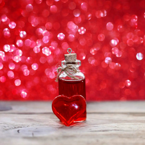Glass bottle with red liquid and cork top next to heart-shaped gemstone on wooden surface against a bokeh red background