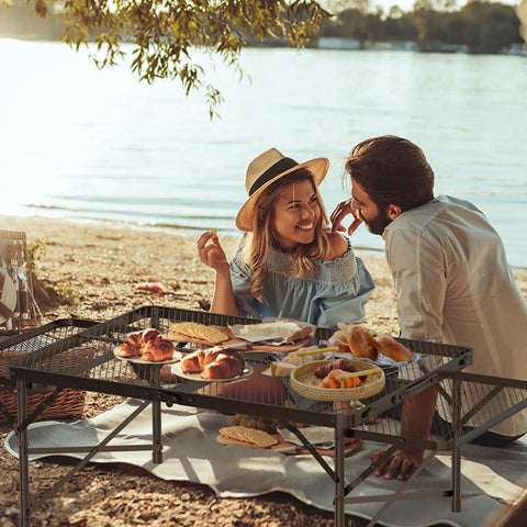 Couple enjoying picnic on beach with tool free foldable outdoor table with wing panels and mesh desktop, 3x2 ft.