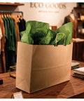 Go-To Heavy-Duty Kraft Paper Gift Bags in brown with reinforced block bottom, standing open with tissue paper on a shop counter, 16x6x13 inches.