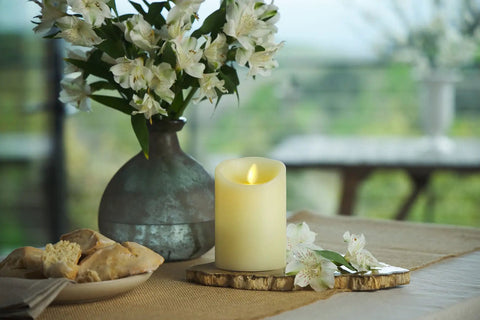Scalloped Edge Flameless LED Candle Ivory 3.5 Inch Diameter 9 Inch Tall Real Wax Pillar With Moving Flame And Auto Timer Battery Operated Vanilla Honey Scented Cozy Glow on table with flowers and bread