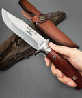 Hand holding M390 Fixed Blade Hunting Knife With Wood Handle Full Tang Survival Knife Leather Sheath Built For Rugged Outdoor Camping Bushcraft And Beast Mode Wilderness Tasks No Cap, showing blade detail and sheath.