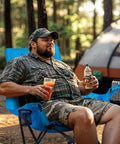 Man sitting on Oversized Rocking Camping Chair with Luxury Padded Recliner in blue, holding drinks, tent in background, outdoor scene